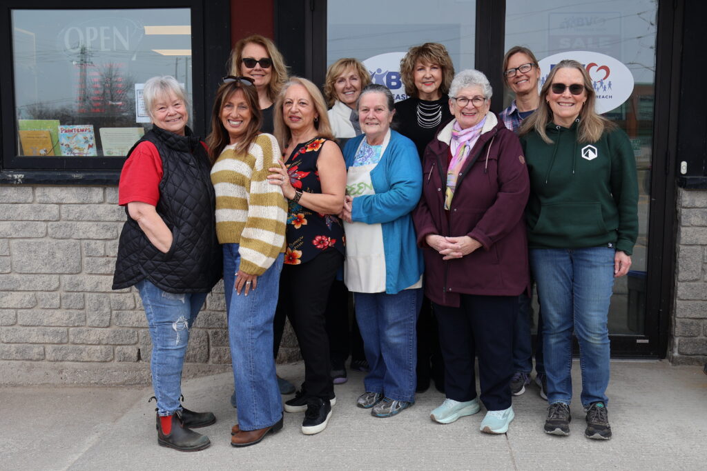 A group of smiling women posing together outside a storefront with glass windows and posters behind them.