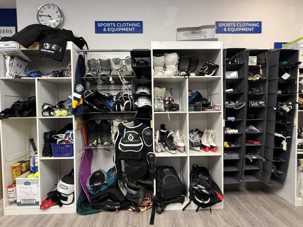 Locker area with cubbies filled with hockey gear: skates, pads, bags, and clothing scattered across shelves under a 'Sports Clothing & Equipment' sign.