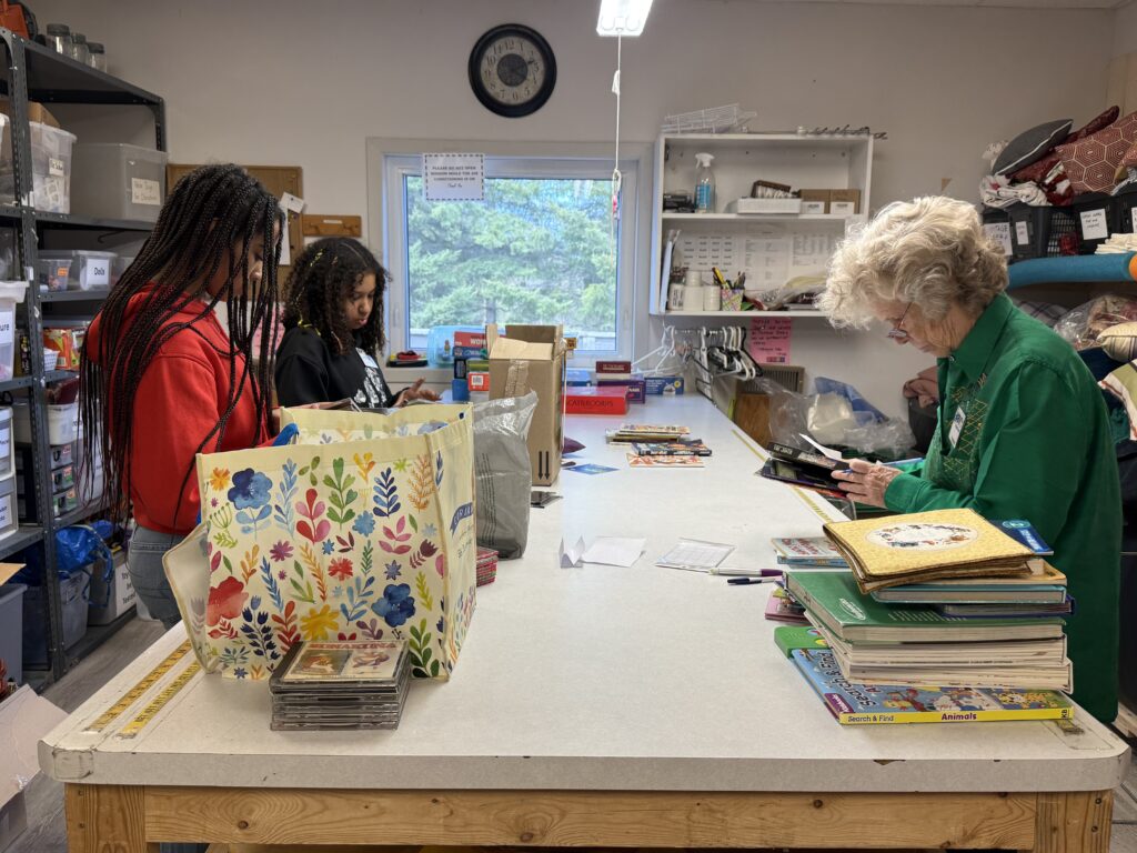 Two young women bag items at a thrift-store checkout while an older woman in a green shirt browses or assists at the counter.