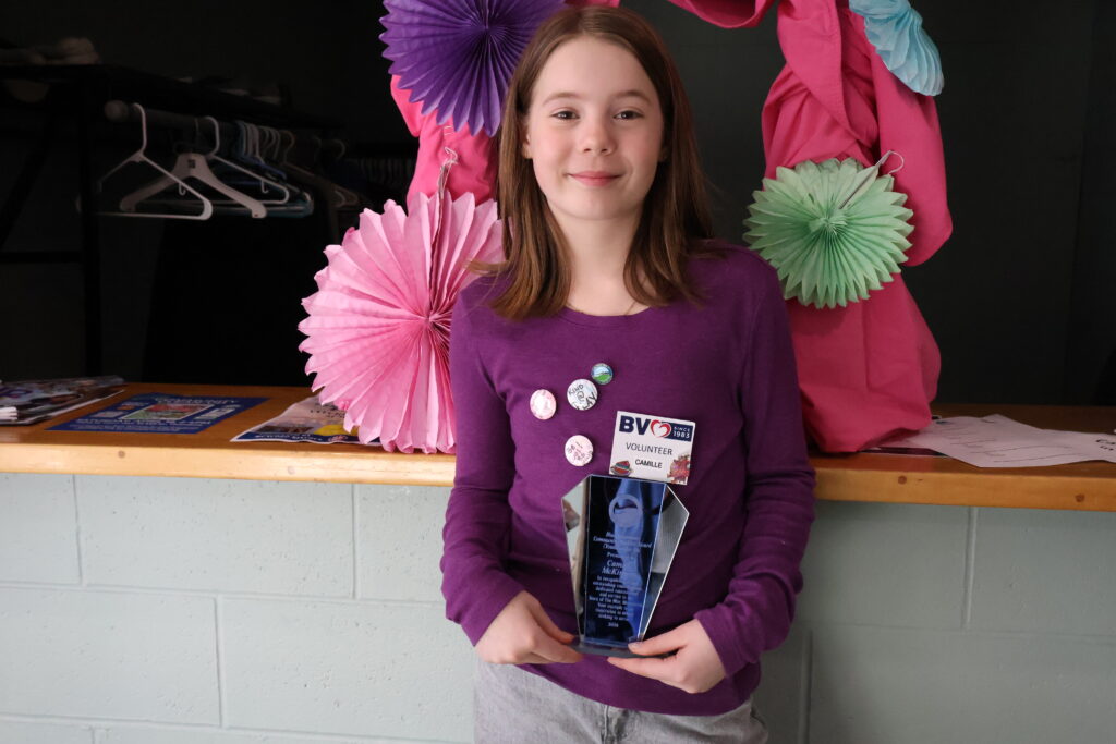 Girl in a purple long-sleeve shirt stands at a table, holding a tall glass trophy with both hands; she wears multiple badges and has colorful paper decorations behind her in pink, purple, and green.