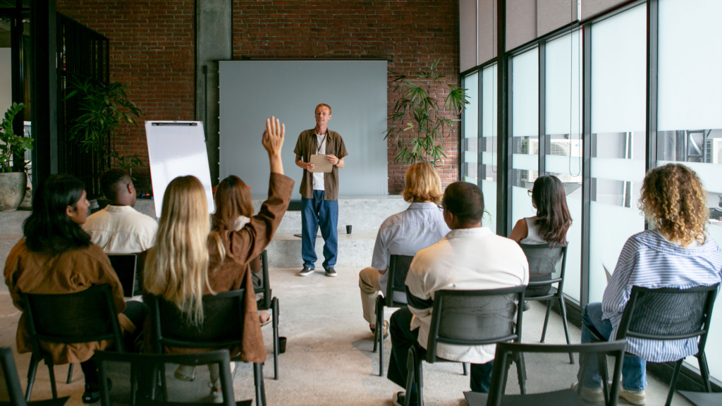 Instructor presenting to a small audience in a brick-walled room with a whiteboard and plants in the background.