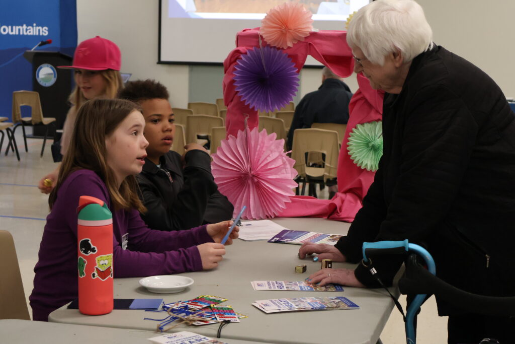 Two girls sit at a craft table with papers and a pink Spongebob water bottle in the foreground, while an adult in a black jacket guides them amid colorful hanging paper decorations.