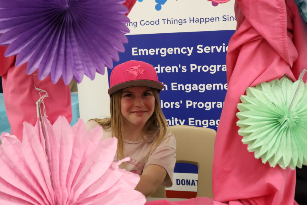 Girl in a pink cap smiles at a photo booth surrounded by bright pink and green paper decorations, at a fundraiser with a 'Donations' box nearby.