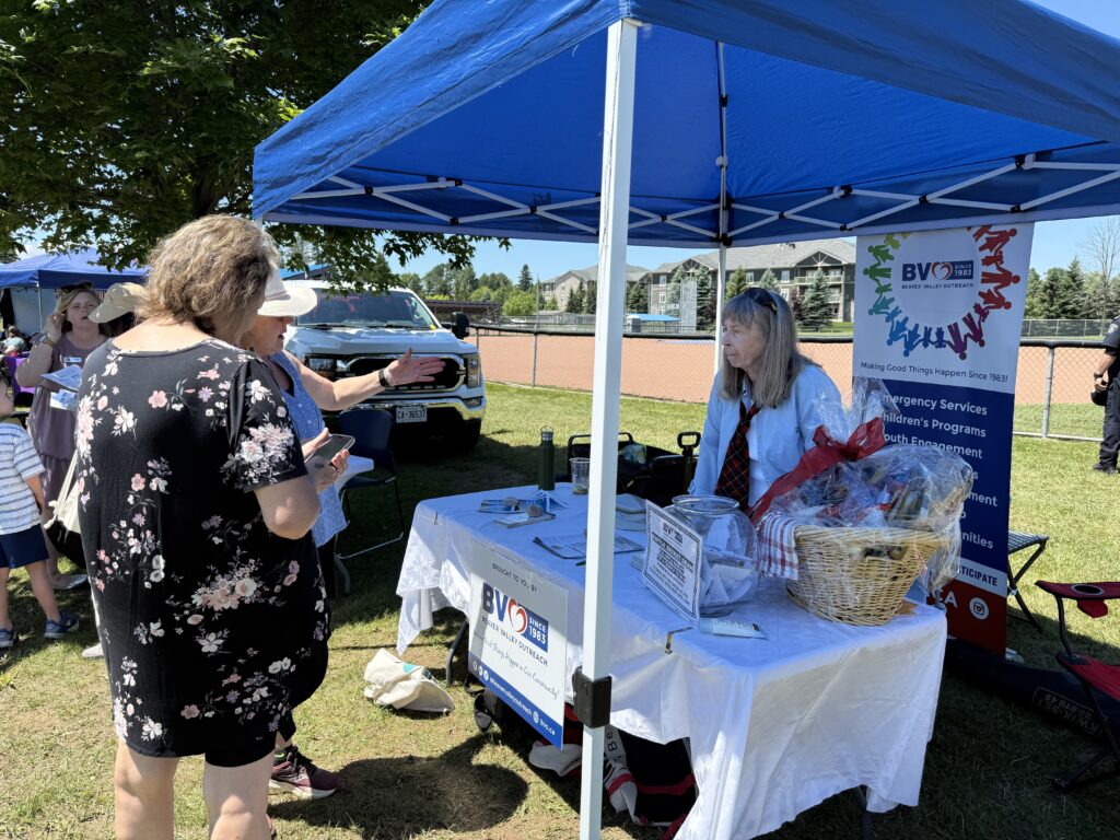 People converse at an outdoor community event under a blue canopy; a banner for Beaver Valley Outreach stands beside a table of materials.