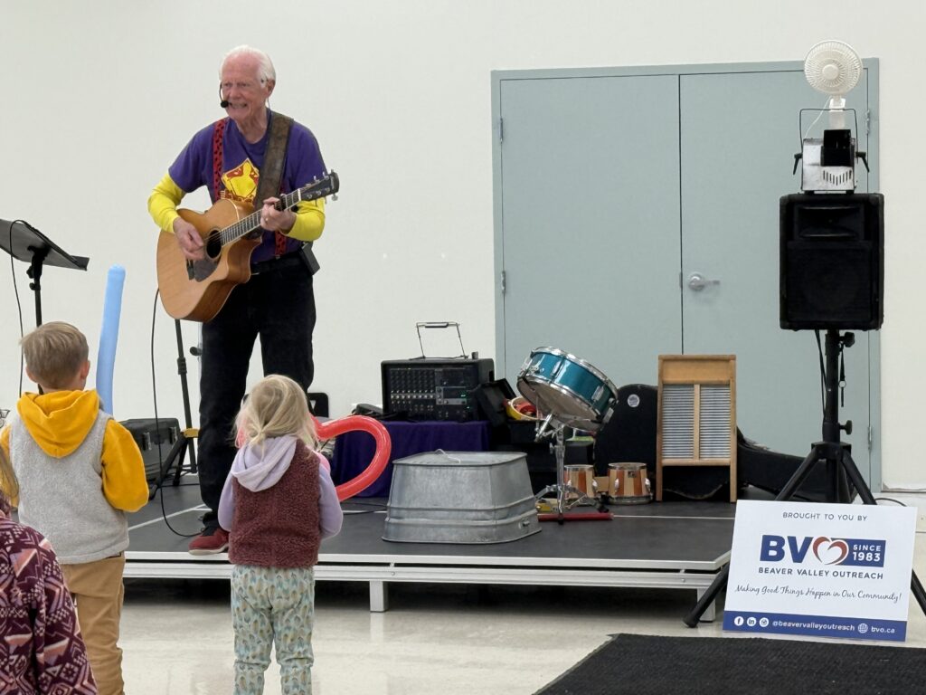 Older man playing an acoustic guitar on a stage while children watch at a community event.