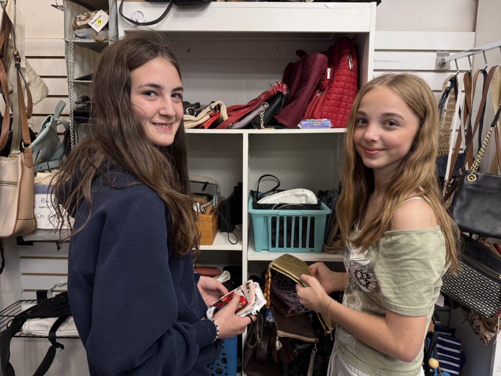 Two young women in a bag shop compare wallets and purses, surrounded by shelves of handbags and accessories.