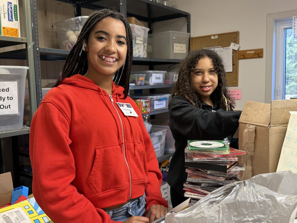 Two smiling volunteers in a storage room with labeled plastic bins; foreground features a girl in a red hoodie with braces, while another girl helps with a cardboard box in the background.