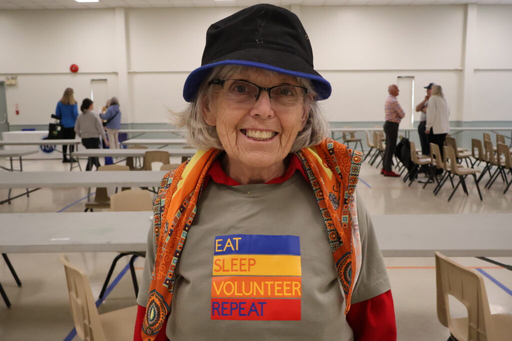 Smiling elderly woman in a black hat with blue trim and a colorful scarf, wearing a gray shirt that reads 'EAT SLEEP VOLUNTEER REPEAT' at an indoor event venue.