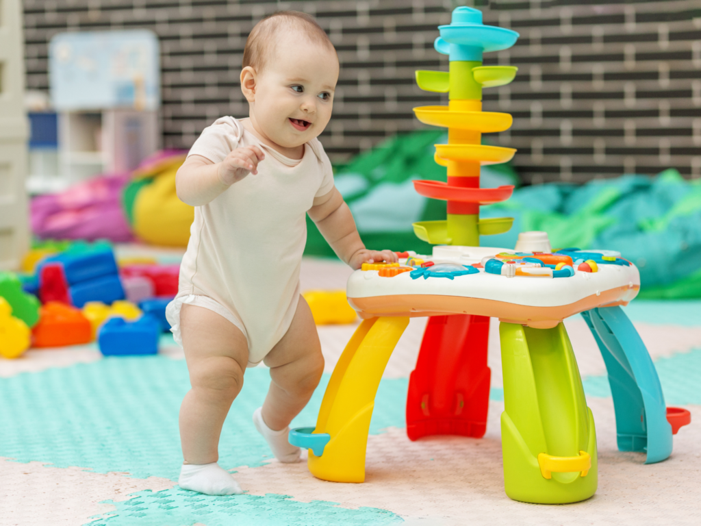 Baby in a light onesie standing at a bright multi‑color activity center, reaching for the next ring, indoors.