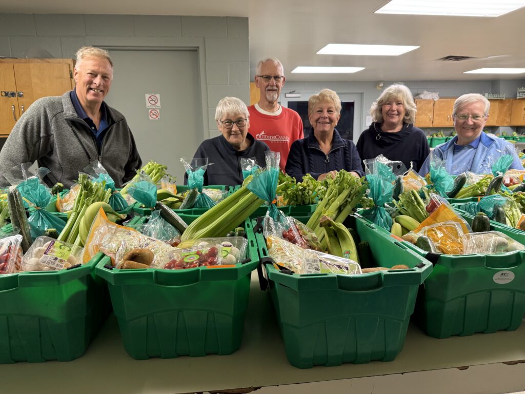 Six volunteers pose behind-green crates of wrapped produce in a food pantry, smiling at the camera.